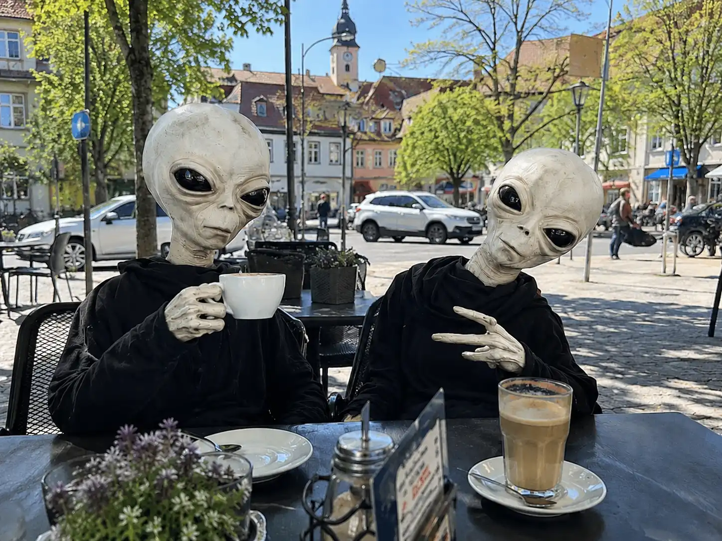 Zwei blassweiße Alienwesen sitzen an einem sonnigen Straßencafé in einer deutschen Kleinstadt und trinken Kaffee zwischen normalen Passanten.